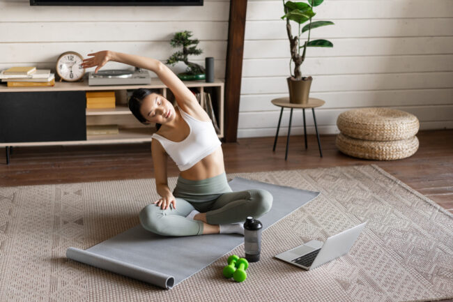 Young asian fitness girl doing stretching, sport workout at home, stretch arms and looking at laptop, follow exercise video on computer